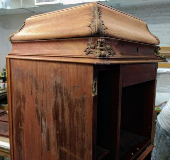 An antique floor-standing Victrola with carved corner details, sun-faded exterior finish, and areas of failing veneer in its original condition before restoration. Antique conservation studio in Chicago, Illinois.